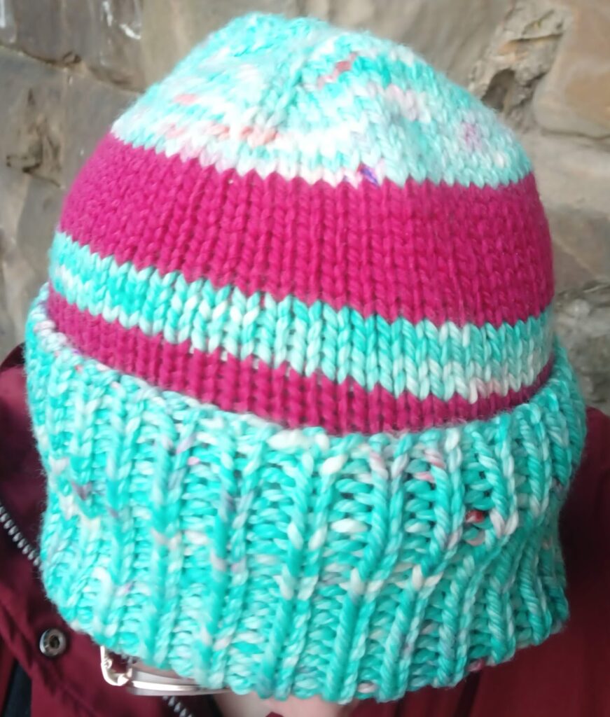A close-up of the top of the aqua Northern Attitude Beanie. You can see the lifted crown increases, and the cerise stripes contrasting with the main aqua yarn. The ribbed brim is thick and cosy.
