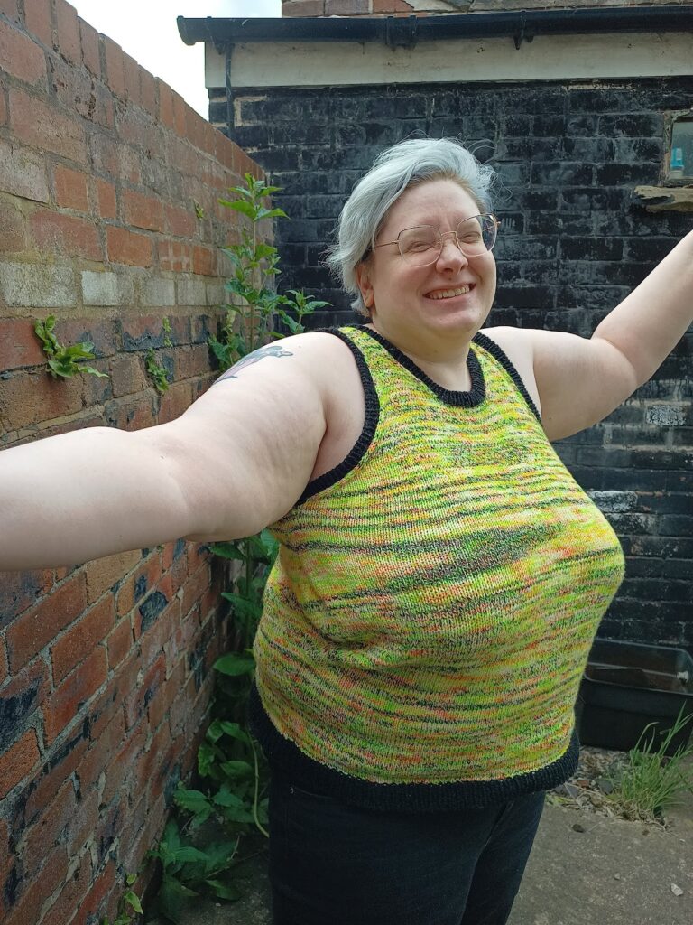 I'm standing in a 3/4 view facing the camera, wearing a brightly coloured, variegated knitted tank top (the Staple Tank by Claire Jackson/Perfectly Knotted) in shades of yellow, green, orange, and pink, with splashes of black. The tank top has black ribbing around the collar, armholes, and bottom hem. My arms are open wide and I am smiling.